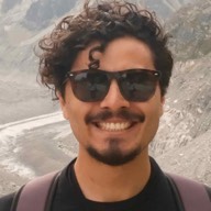 A headshot of Julian wearing sun glasses. Short, curly hair, cinnamon-coloured skin, moustache and goatee. Big smile